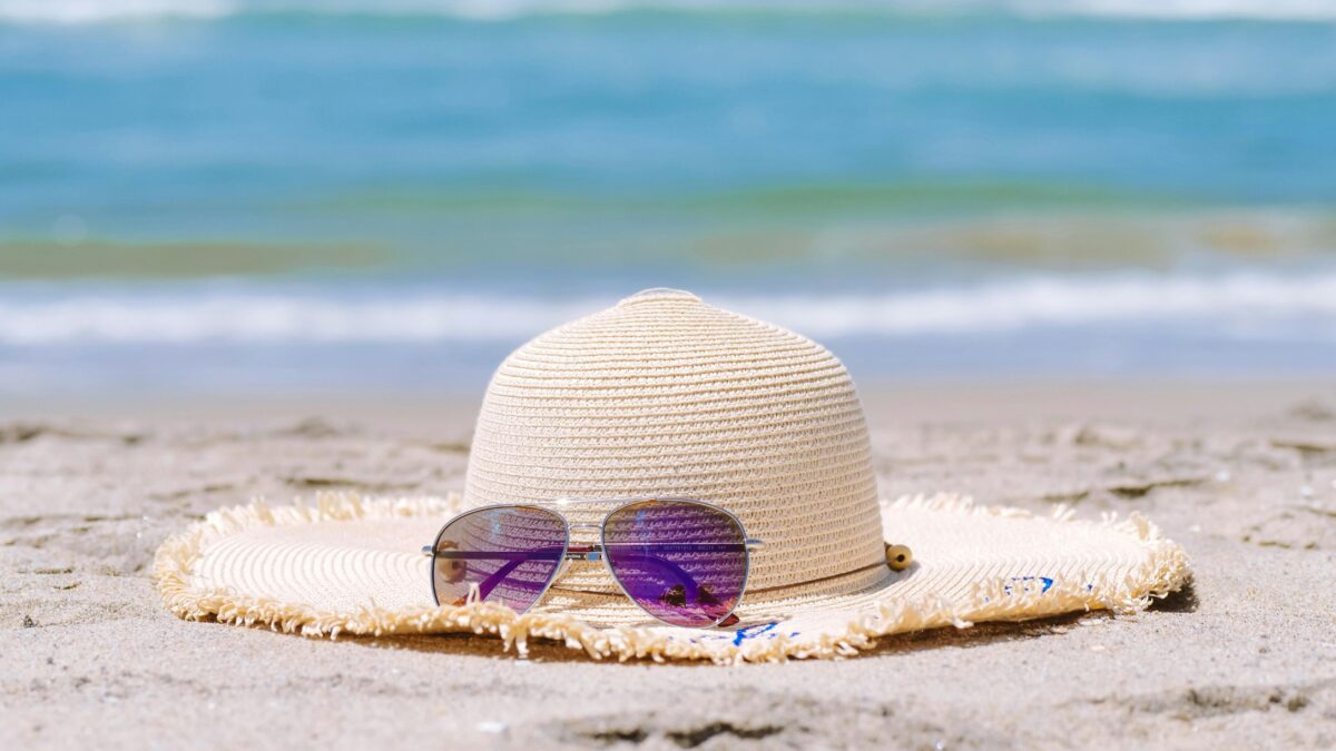 Ein Strohhut und eine Sonnenbrille, die auf dem Strand liegen. Im Hintergrund sieht man blaues Wasser.